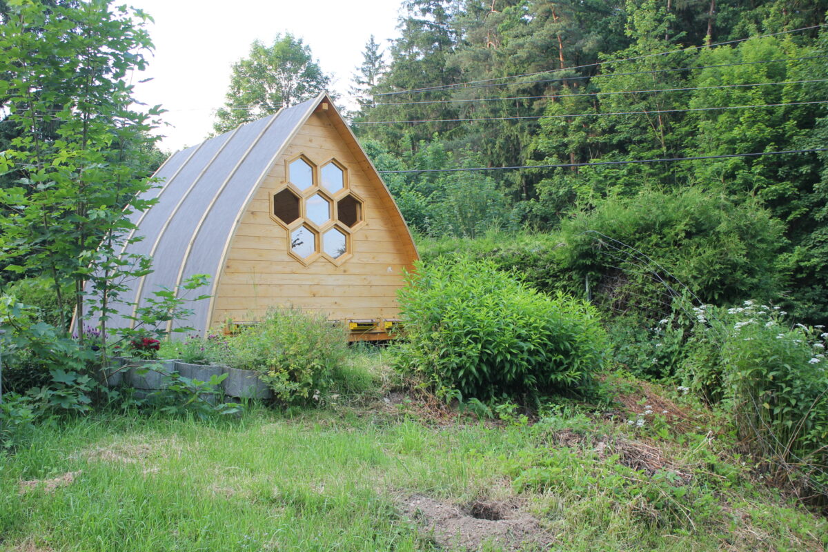 Bienenstockluft-Hütte aus Holz mit hexagonalen Wabenfenstern in grüner Gartenlandschaft im Mühlviertel